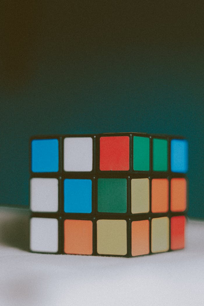 Unsolved three dimensional magic cube with multicolored stickers placed on white table against dark wall