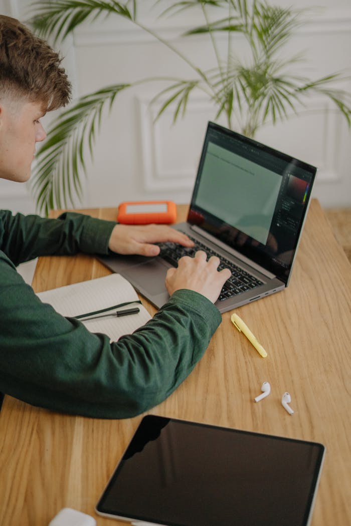 A young man using a laptop for online learning at a wooden desk with plants indoors.